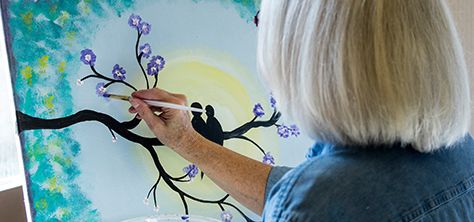 An elderly woman paints purple flowers on a tree